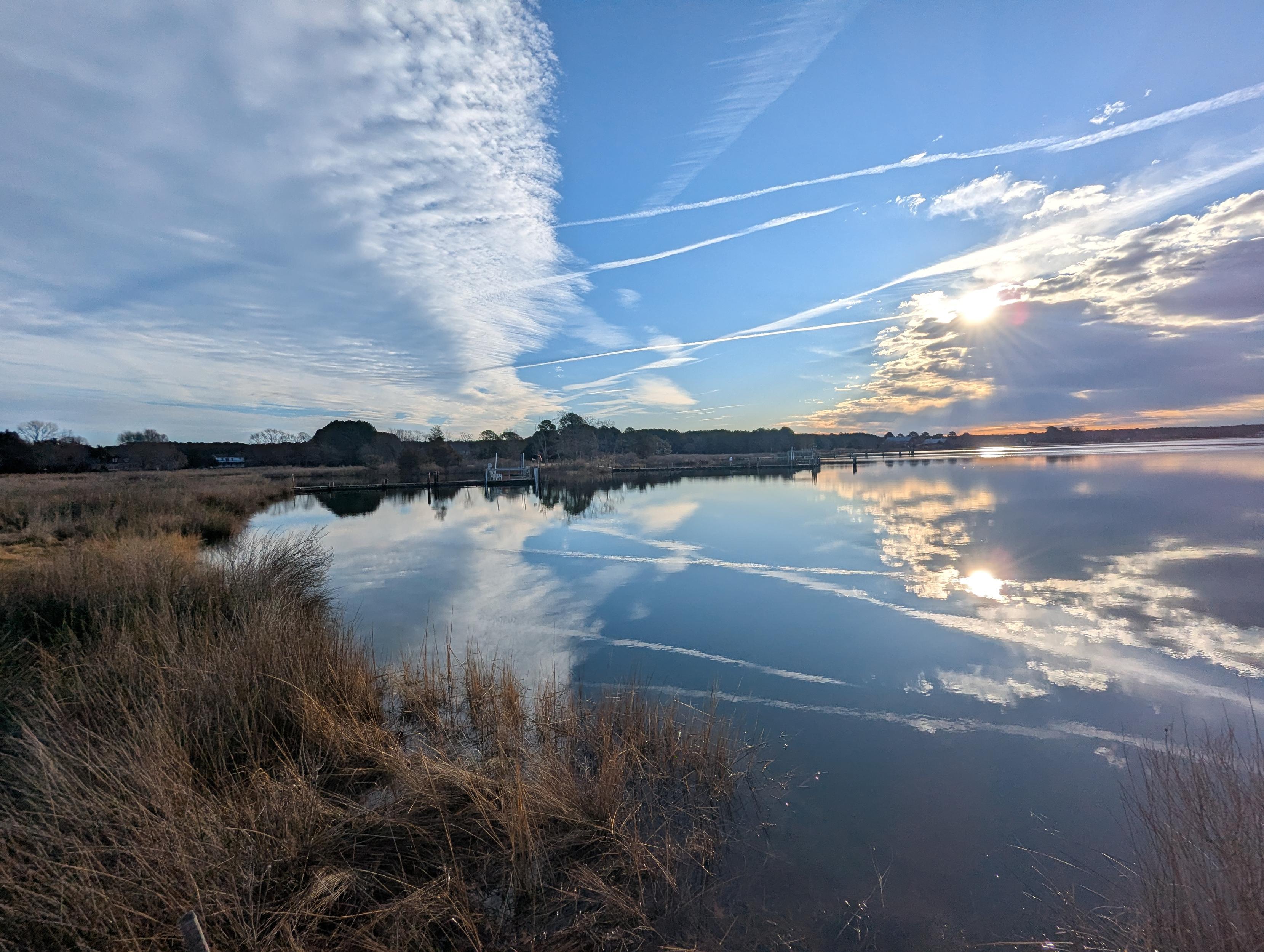 Photo of a calm morning here on Harris Creek. Sun rising low  to the right in a deep blue sky behind a long cloud bank. Marsh is brown and flooded with the high tide. Creek water is rippled and reflects the sun, sky and clouds.