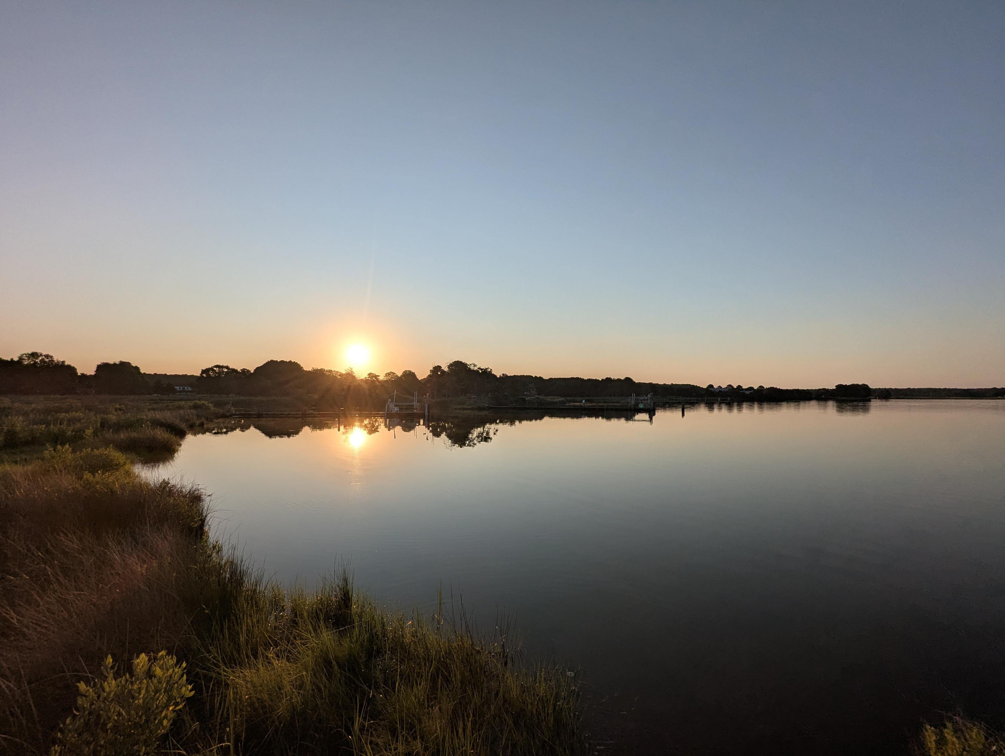Photo of a calm morning sunrise over Harris Creek. Sun rising low to the left in a clear blue sky. Marsh is brilliantly green and flooded. Creek water is rippled and reflects the sun and sky.