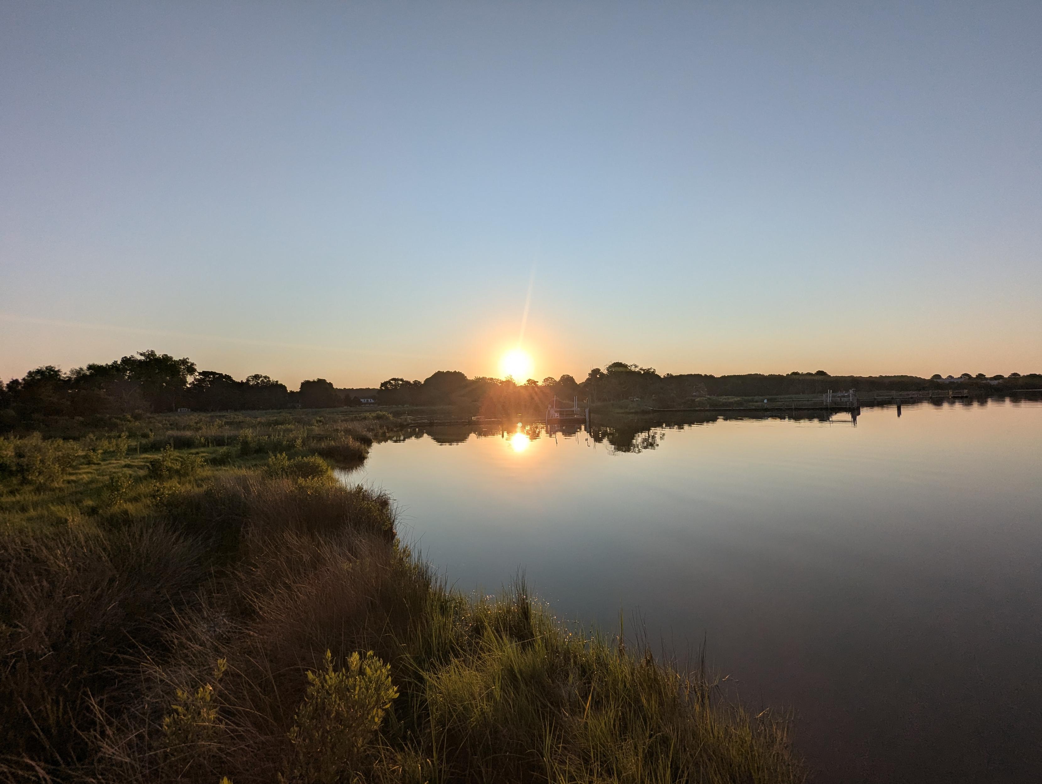 Photo of a calm morning sunrise over Harris Creek. Sun rising low to the left in a clear blue sky. Marsh is brilliantly green and flooded. Creek water is rippled and reflects the sun and sky.