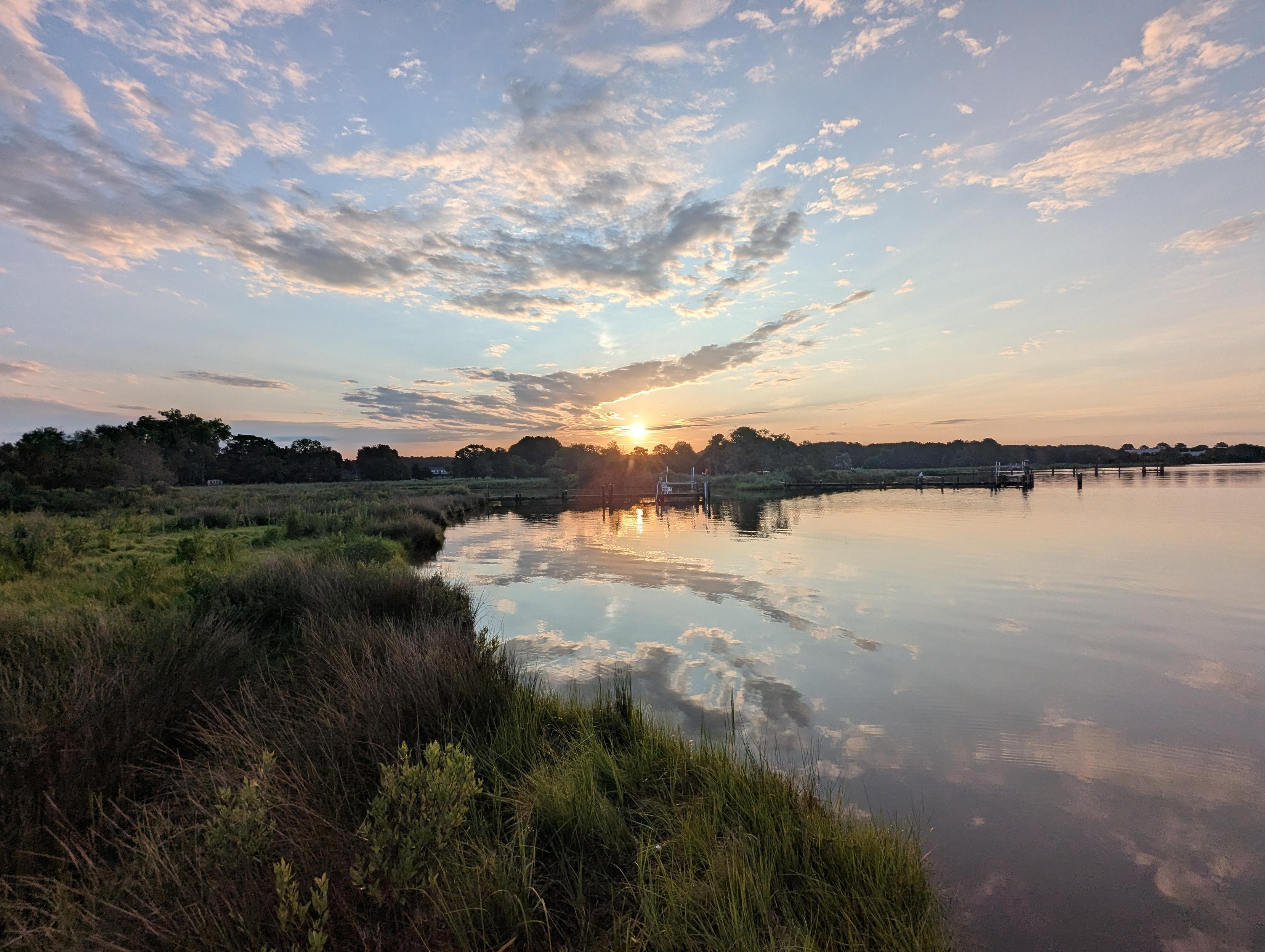 Photo of a calm morning sunrise over Harris Creek. Sun rising low to the left in a pale blue sky. The horizon is lit orange. Scattered mid level clouds ride the sky.