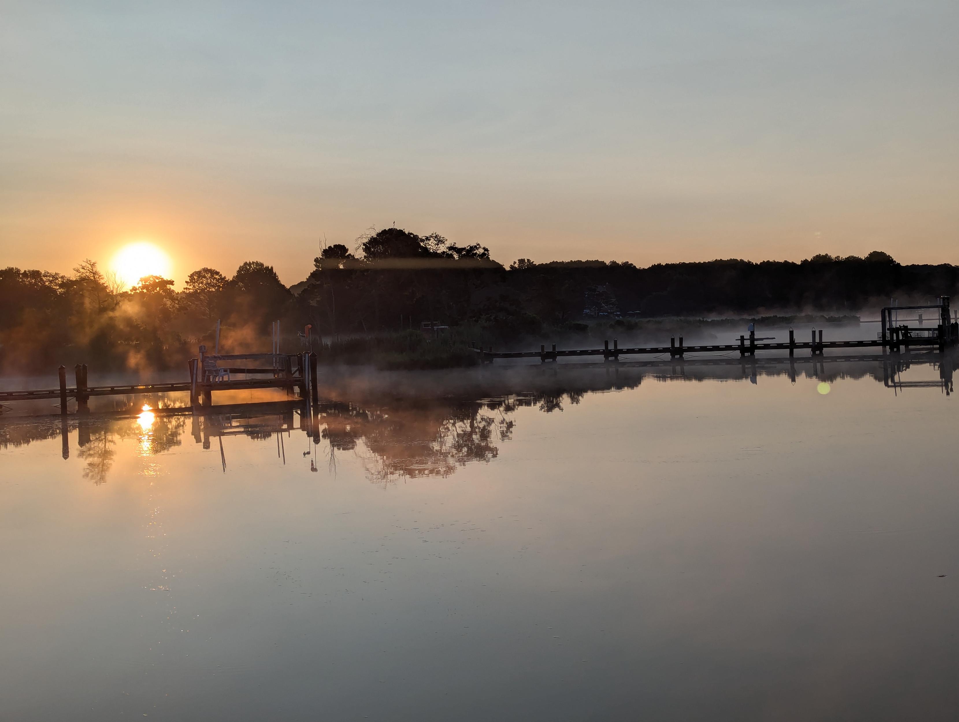Photo of a calm morning sunrise over Harris Creek. Sun rising low to the left just over the horizon. Sky is pale blue and slightly hazy. Wispy fog rises close from the water. Marsh is brilliantly green and wet with the morning dew. Creek water is rippled and reflects the sun and sky.