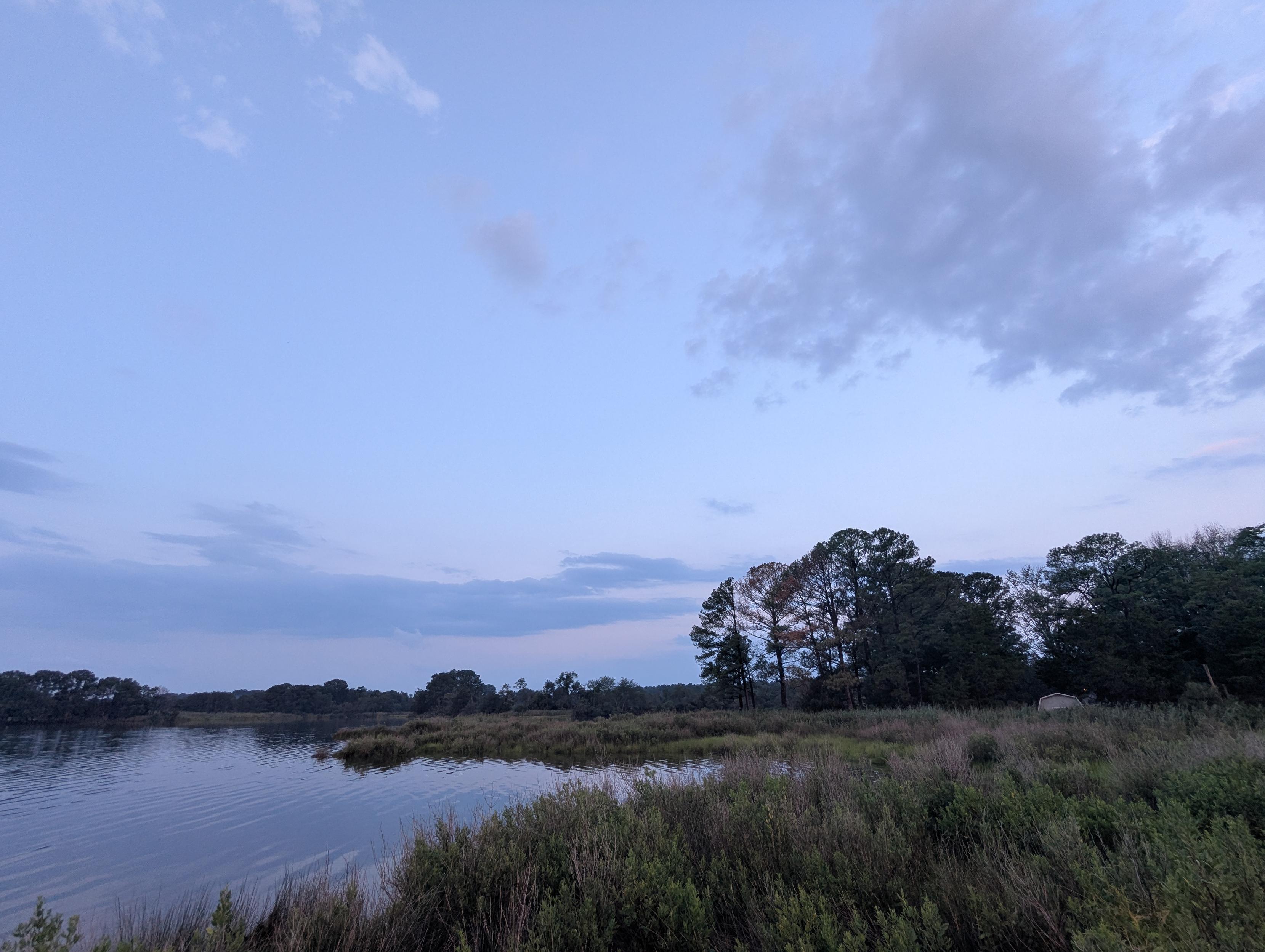 Photo of a dramatic cloudscape in a post-storm morning sunrise here on Harris Creek.
Sun rising low to the left behind a dark building cumulus cloud. Sky is decorated with  multilevel grey clouds, lit orange in the East by the rising sun. Marsh is brilliantly green and flooded. Creek water is rippled and reflects the sun, sky and clouds.
