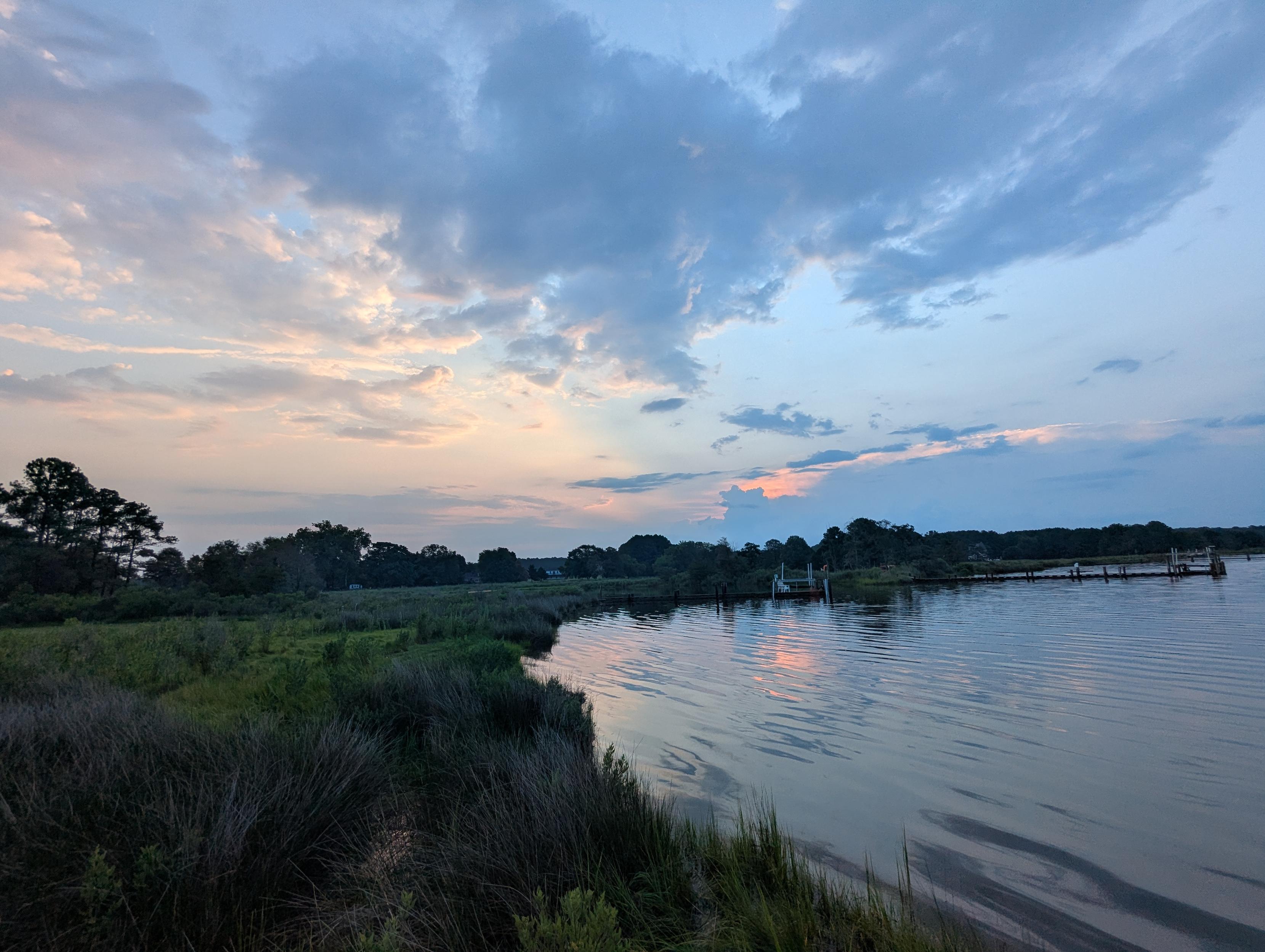 Photo of a dramatic cloudscape in a post-storm morning sunrise here on Harris Creek.
Sun rising low to the left behind a dark building cumulus cloud. Sky is decorated with  multilevel grey clouds, lit orange in the East by the rising sun. Marsh is brilliantly green and flooded. Creek water is rippled and reflects the sun, sky and clouds.